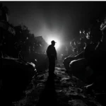 Silhouette of a scrapyard worker standing between piles of crushed cars with a bright light in the background.