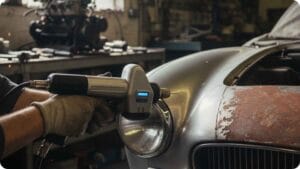 Mechanic using a handheld laser cleaning tool on a vintage classic car’s rusted fender inside an automotive restoration workshop.